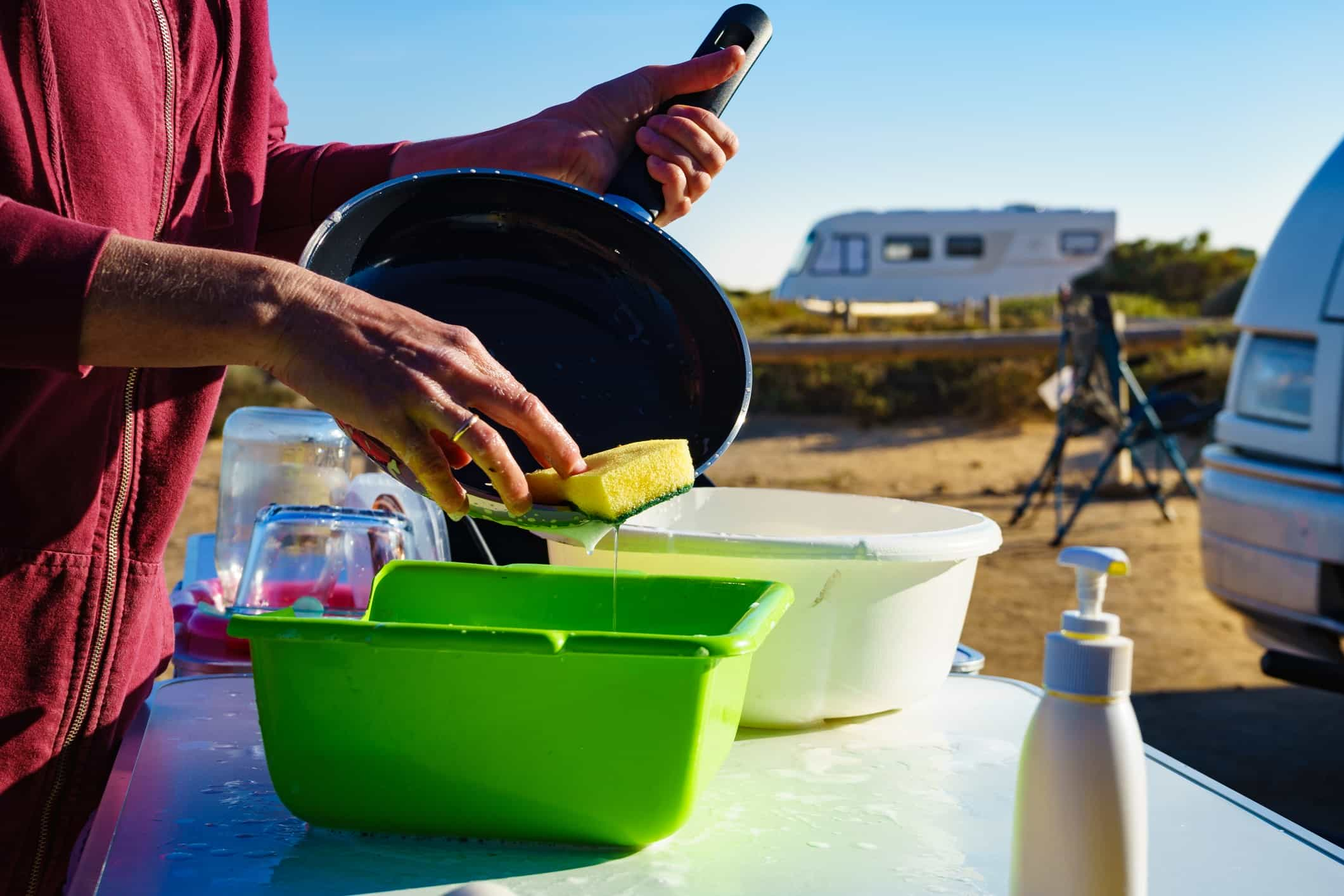 Mature woman washing up dishes in bowl on fresh air. Dishwashing outdoor on camping site, sea shore.