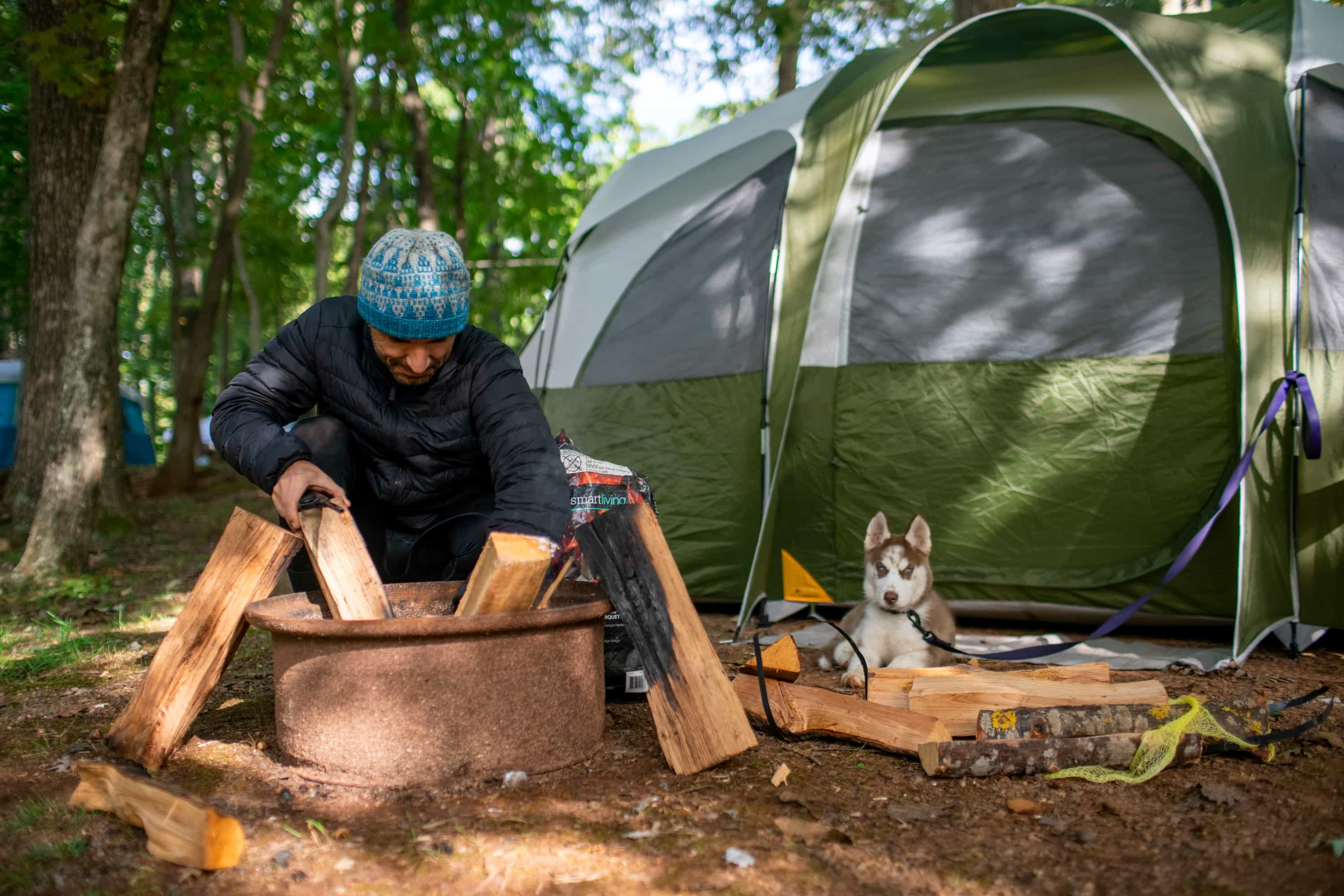 Man building a campfire in front of his tent at KOA with a puppy nearby.